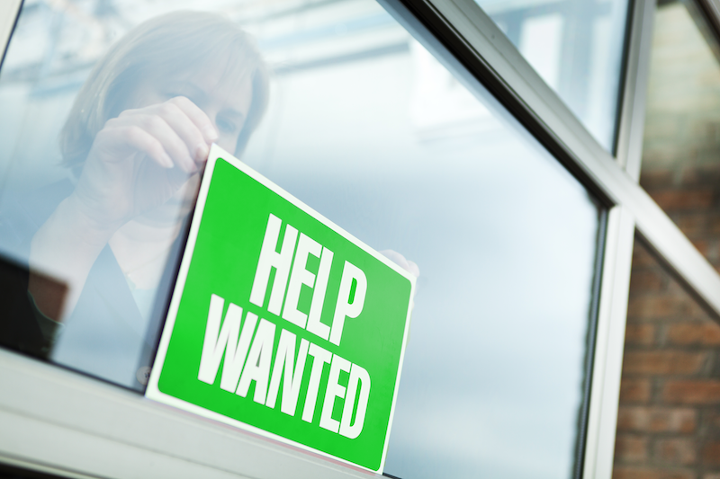 Woman hangs a green "Help Wanted" sign in a business window.
