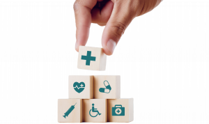 Hand reaching down from out of frame, placing a building block with a Healthcare Plus icon on top; placing the block on a stack of other blocks with various insurance related icons.